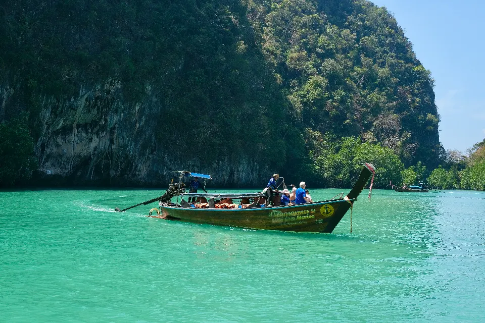 A longtail boat crossing turquoise water below a forested cliff.